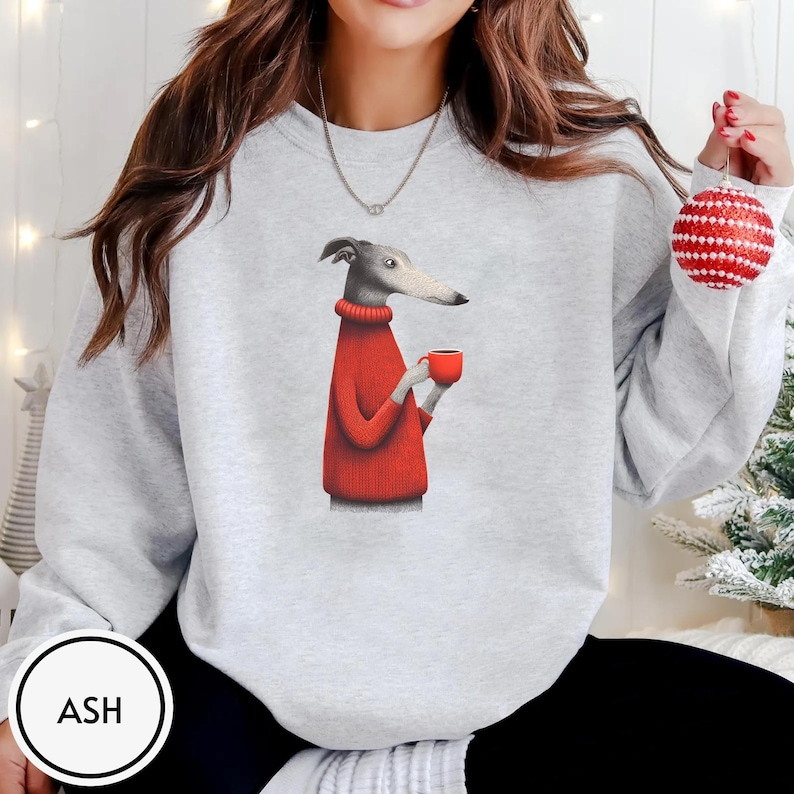 May include: Light gray sweatshirt featuring a whimsical illustration of a greyhound dog wearing a red sweater and holding a red mug. The dog is depicted in a cartoon style. The sweatshirt has a crew neck and long sleeves. The word "ASH" is in a white circle.