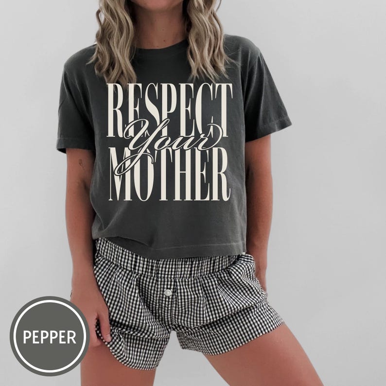 May include: A charcoal gray short-sleeve t-shirt featuring the white text "RESPECT YOUR MOTHER" in a mix of serif and script fonts, paired with black and white gingham patterned shorts that have a single button detail at the center waist.