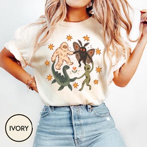 May include: An ivory short-sleeve t-shirt featuring a graphic print of a Bigfoot, a moth-like creature, a green dinosaur, and a green alien holding hands in a circle, surrounded by orange stars and a small flower in the center of the design.
