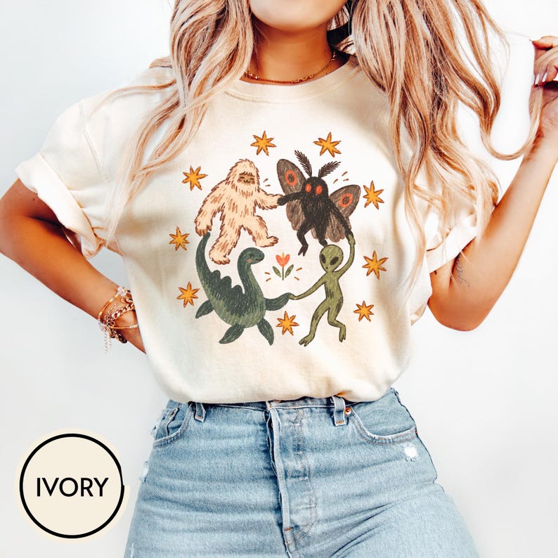 May include: An ivory short-sleeve t-shirt featuring a graphic print of a Bigfoot, a moth-like creature, a green dinosaur, and a green alien holding hands in a circle, surrounded by orange stars and a small flower in the center of the design.