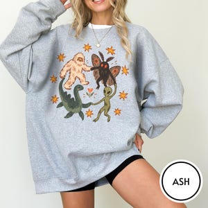 May include: An ash gray long-sleeve sweatshirt featuring a graphic print of a beige yeti, a black moth, a green dinosaur, and a green alien holding hands in a circle. The design includes orange stars and a small flower, all centered on the chest.