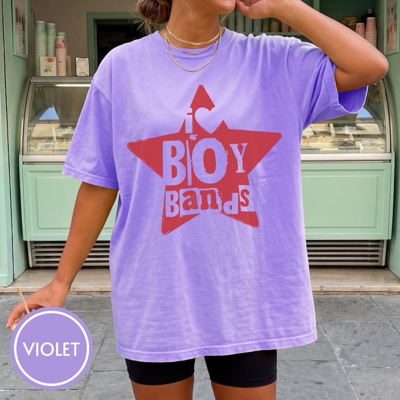 Puede incluir: Una camiseta de manga corta de color violeta claro con un gr&aacute;fico de una estrella roja grande en el pecho. Dentro de la estrella, el texto dice I s&iacute;mbolo de coraz&oacute;n BOY BANDS en una fuente negrita y desgastada. Tiene cuello redondo y corte holgado.