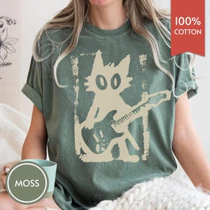 Puede incluir: Camiseta verde con un gr&aacute;fico desgastado de un gato tocando la guitarra el&eacute;ctrica. El gato es beige y la camiseta tiene el texto "100% Algod&oacute;n" en una etiqueta roja. Una taza con la palabra "MOSS" est&aacute; en primer plano.