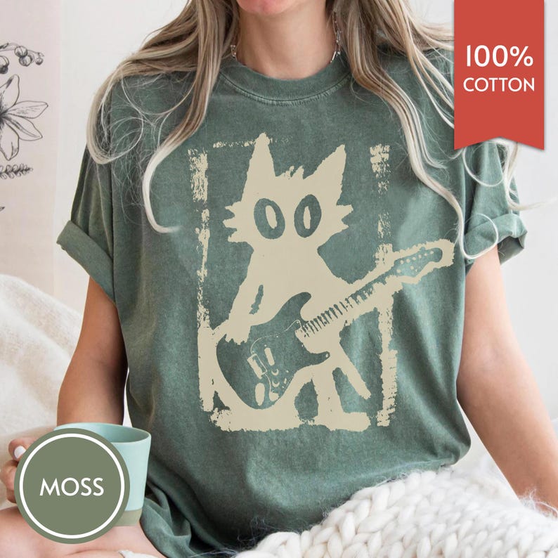 Puede incluir: Camiseta verde con un gr&aacute;fico desgastado de un gato tocando la guitarra el&eacute;ctrica. El gato es beige y la camiseta tiene el texto "100% Algod&oacute;n" en una etiqueta roja. Una taza con la palabra "MOSS" est&aacute; en primer plano.