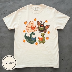 Puede incluir: Una camiseta de manga corta color marfil con un estampado gráfico que muestra a un yeti blanco, una criatura similar a una polilla, un dinosaurio verde y un alienígena verde, cada uno leyendo un libro. El diseño está rodeado de pequeñas estrellas naranjas.