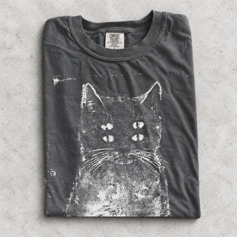 May include: A charcoal grey t-shirt featuring a white screen-printed graphic of a cat with four eyes. The shirt has a crew neckline and a Comfort Colors brand label printed on the inside collar area, indicating size L and made in Honduras.