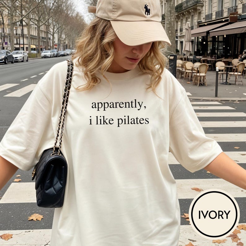 May include: Ivory-colored t-shirt with the text "apparently, i like pilates" in black. The shirt is paired with a beige baseball cap and a black purse with a chain strap. The shirt has short sleeves.