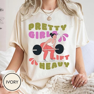 Puede incluir: Camiseta color marfil con el texto "PRETTY GIRLS LIFT HEAVY" en verde y rosa. El gráfico muestra a una mujer levantando pesas. La camiseta es de estilo cómodo e informal.