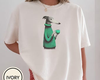 May include: An ivory short-sleeved t-shirt featuring a graphic illustration of a greyhound dog wearing a green ribbed sweater and holding a small green mug. The dog is depicted in profile, facing right, with a neutral expression on its face.