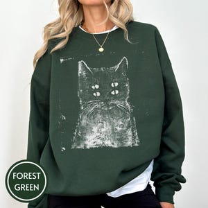 May include: A forest green crewneck sweatshirt featuring a white, distressed-style graphic print of a cat with four eyes. The design has a textured, ink-stamped appearance centred on the chest. The garment has long sleeves and a ribbed neckline.