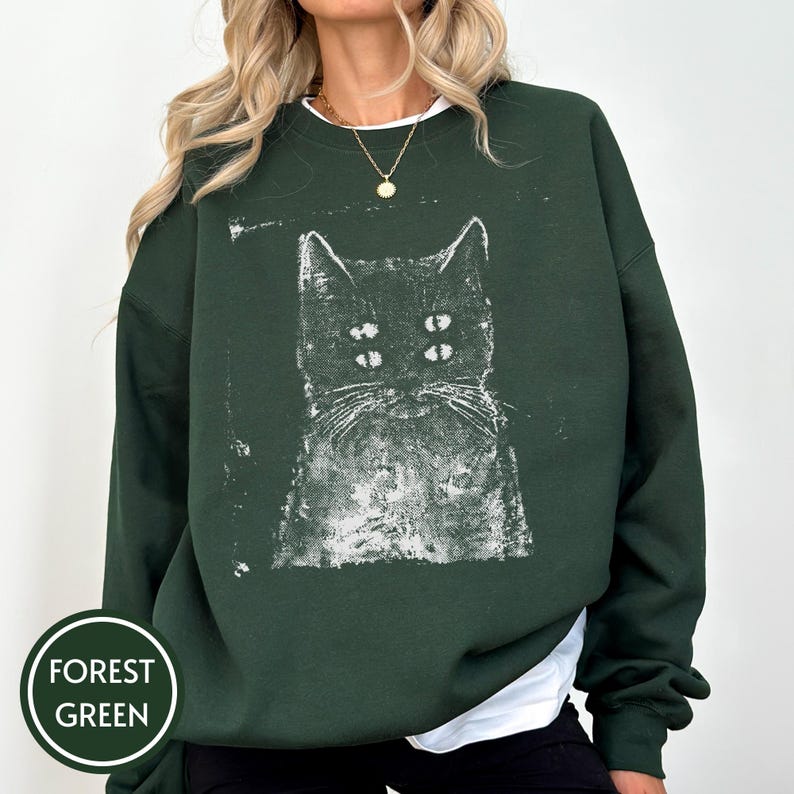 May include: A forest green crewneck sweatshirt featuring a white, distressed-style graphic print of a cat with four eyes. The design has a textured, ink-stamped appearance centred on the chest. The garment has long sleeves and a ribbed neckline.