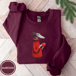 May include: Maroon sweatshirt featuring a graphic of a greyhound dog wearing a red sweater and holding a red mug. The sweatshirt has a crew neck and long sleeves. The word "MAROON" is printed in a white circle at the bottom.