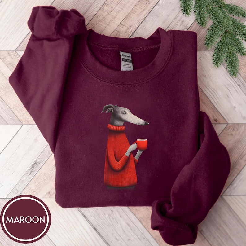 May include: Maroon sweatshirt featuring a graphic of a greyhound dog wearing a red sweater and holding a red mug. The sweatshirt has a crew neck and long sleeves. The word "MAROON" is printed in a white circle at the bottom.