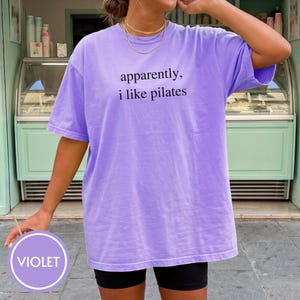 May include: A violet-colored t-shirt with the text "apparently, i like pilates" in black. The shirt is a relaxed fit and has short sleeves. The image also includes a violet-colored circle with the word "VIOLET" in white.