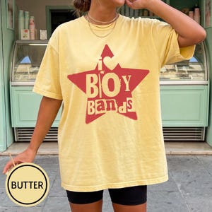 Puede incluir: Una camiseta de manga corta de color amarillo p&aacute;lido con un gr&aacute;fico de una estrella roja grande en el pecho. Dentro de la estrella, el texto dice I heart BOY BANDS en una fuente negrita y desgastada. La camiseta tiene cuello redondo y corte holgado.