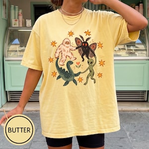 May include: A butter yellow short-sleeve t-shirt featuring a central graphic of a white yeti, a black mothman, a green sea monster, and a green alien holding hands in a circle, surrounded by orange stars and a small flower in the center.