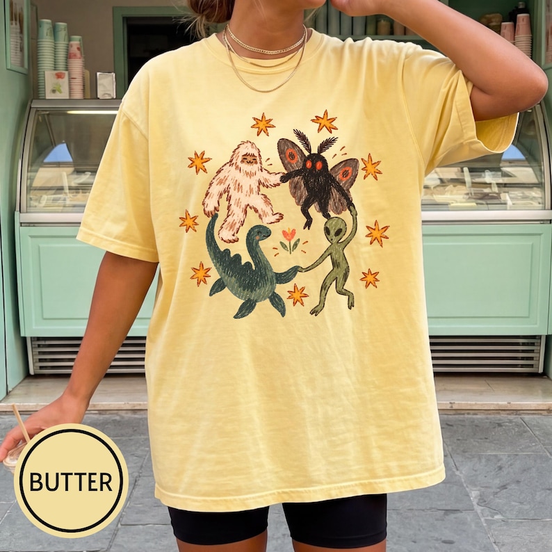 May include: A butter yellow short-sleeve t-shirt featuring a central graphic of a white yeti, a black mothman, a green sea monster, and a green alien holding hands in a circle, surrounded by orange stars and a small flower in the center.