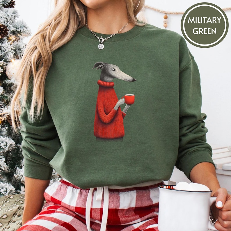 May include: Military green sweatshirt featuring a graphic of a greyhound wearing a red sweater and holding a red mug. The sweatshirt has long sleeves and a crew neck. The text "MILITARY GREEN" is in a circle on the upper right.