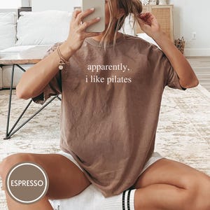May include: Espresso-colored t-shirt with the text "apparently, i like pilates" in white. The shirt is a relaxed fit and appears to be made of soft material. The image shows the shirt being worn, suggesting it's part of a casual wear collection.