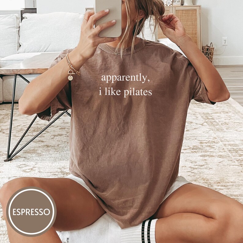 May include: Espresso-colored t-shirt with the text "apparently, i like pilates" in white. The shirt is a relaxed fit and appears to be made of soft material. The image shows the shirt being worn, suggesting it's part of a casual wear collection.