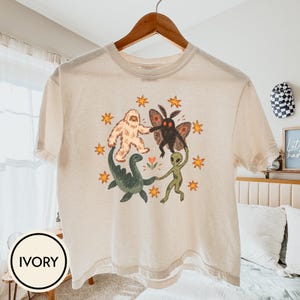 Peut inclure: Un t-shirt à manches courtes ivoire avec une impression graphique circulaire représentant un yéti blanc, une créature papillon noire, un monstre marin vert et un extraterrestre vert se tenant la main, entourés d'étoiles orange et d'une fleur.