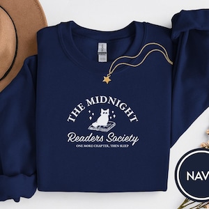 Op de afbeelding: Marineblauwe sweatshirt met de tekst "The Midnight Readers Society" en een witte afbeelding van een kat op een boek. Een gouden ketting met een sterhanger ligt op de sweatshirt. Een bruine hoed en bloemen zijn op de achtergrond.