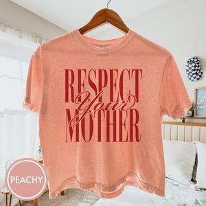 May include: A peach-colored short-sleeve t-shirt featuring the text "RESPECT YOUR MOTHER" printed in a bold, dark red font. The shirt has a crew neckline and a standard fit, displayed on a wooden hanger against a light-colored bedroom interior.