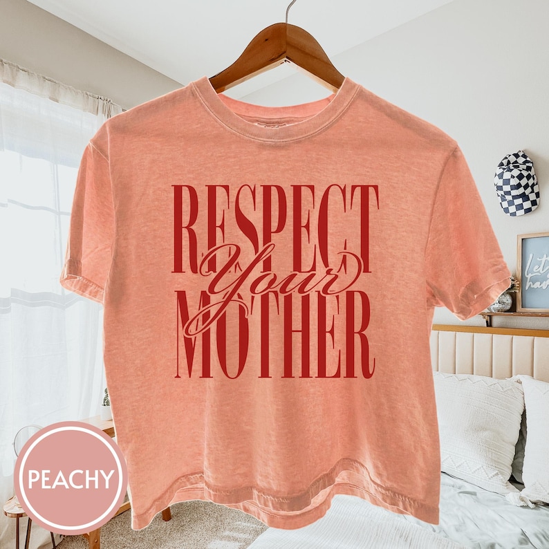 May include: A peach-colored short-sleeve t-shirt featuring the text "RESPECT YOUR MOTHER" printed in a bold, dark red font. The shirt has a crew neckline and a standard fit, displayed on a wooden hanger against a light-colored bedroom interior.