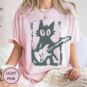 Puede incluir: Camiseta rosa claro con un gato gris tocando la guitarra. El gr&aacute;fico del gato est&aacute; dentro de un marco rectangular. Las palabras "LIGHT PINK" est&aacute;n en un c&iacute;rculo en la esquina inferior izquierda. La camiseta tiene mangas cortas.