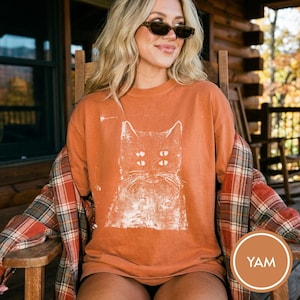 May include: A burnt orange crewneck t-shirt featuring a distressed white graphic print of a cat with four eyes. The shirt is layered under a red, white, and grey plaid flannel button-down shirt with long sleeves and a classic collar design.