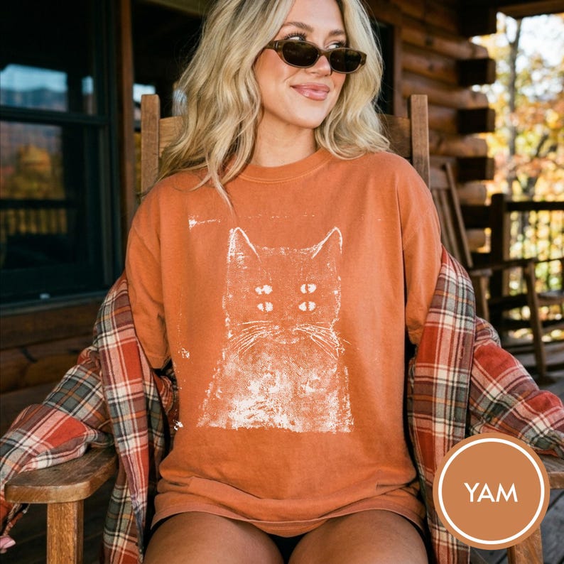 May include: A burnt orange crewneck t-shirt featuring a distressed white graphic print of a cat with four eyes. The shirt is layered under a red, white, and grey plaid flannel button-down shirt with long sleeves and a classic collar design.