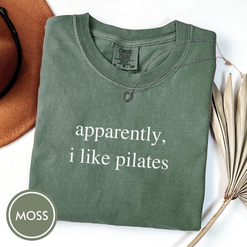 May include: A moss green t-shirt with the text "apparently, i like pilates" in white. A silver necklace with a pendant is draped on the shirt. The shirt is folded on a white surface.
