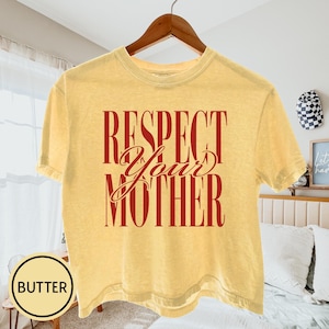 May include: A butter yellow short-sleeve t-shirt featuring the dark red text "RESPECT YOUR MOTHER" printed in a bold, vertical block font with the word "Your" in a cursive script style across the center of the chest.