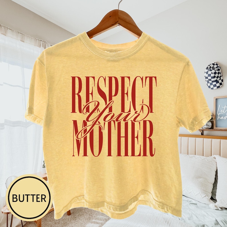 May include: A butter yellow short-sleeve t-shirt featuring the dark red text "RESPECT YOUR MOTHER" printed in a bold, vertical block font with the word "Your" in a cursive script style across the center of the chest.