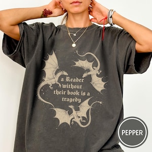Op de afbeelding: Een antracietgrijs T-shirt met korte mouwen en een grafische print van drie draken rondom de tekst "a Reader without their book is a tragedy". Het shirt heeft een vervaagde vintage look en een ronde hals. Een rond label bevat de tekst "PEPPER".