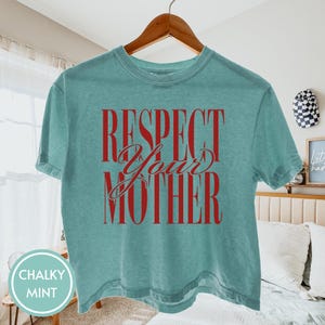 May include: A chalky mint green short-sleeve t-shirt featuring the red text "RESPECT YOUR MOTHER" printed in a combination of bold block lettering and a cursive script font across the center of the chest, hanging on a wooden hanger.