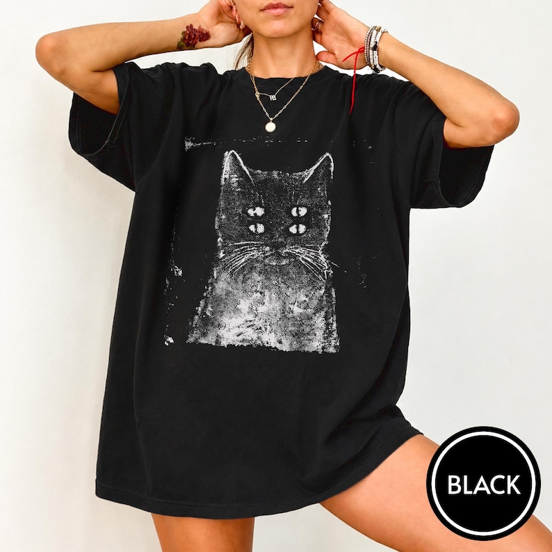 May include: A black short-sleeved t-shirt featuring a white graphic print of a cat with four eyes. The design has a distressed, textured appearance. The shirt has a crew neckline and a loose, relaxed fit, providing a casual look for everyday wear.