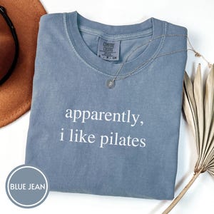 May include: A blue jean colored t-shirt with the text "apparently, i like pilates" in white. A silver necklace with a small pendant rests on the shirt. The shirt is folded neatly on a white surface.