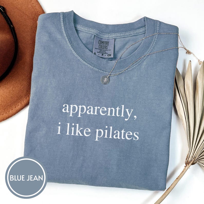 May include: A blue jean colored t-shirt with the text "apparently, i like pilates" in white. A silver necklace with a small pendant rests on the shirt. The shirt is folded neatly on a white surface.