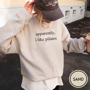 May include: Sand-colored sweatshirt with the text "apparently, i like pilates" in black. The sweatshirt is paired with a white undershirt and a brown baseball cap. A gold necklace is visible. The image has a neutral, casual aesthetic.