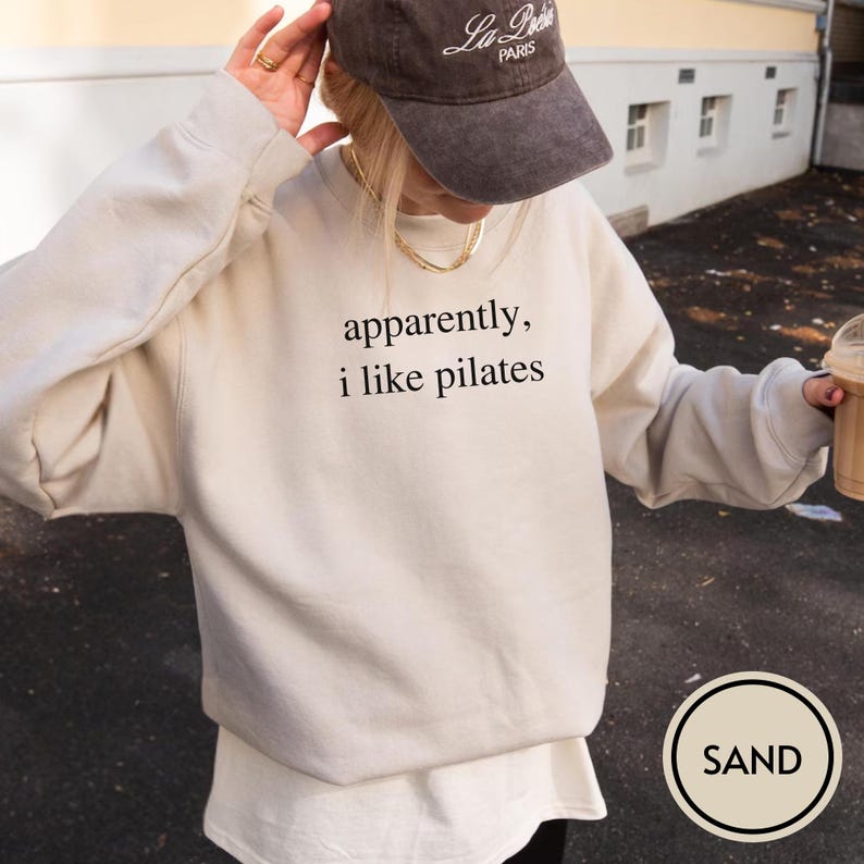 May include: Sand-colored sweatshirt with the text "apparently, i like pilates" in black. The sweatshirt is paired with a white undershirt and a brown baseball cap. A gold necklace is visible. The image has a neutral, casual aesthetic.
