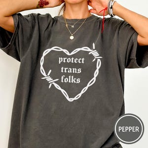 Op de afbeelding: Donkergrijs t-shirt met een wit hartvormig ontwerp van prikkeldraad. De woorden "protect trans folks" zijn in gotische letters in het midden van het hart gedrukt. Het shirt heeft korte mouwen en een ronde hals.