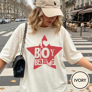 Puede incluir: Una camiseta de manga corta color marfil con un gr&aacute;fico de una estrella roja grande en la parte delantera. Dentro de la estrella, el texto dice I s&iacute;mbolo de coraz&oacute;n BOY BANDS en una fuente blanca desgastada. Tiene cuello redondo y corte holgado.
