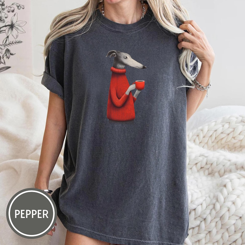May include: Dark gray t-shirt featuring a cartoon greyhound dog wearing a red sweater and holding a red cup. The shirt has short sleeves and a relaxed fit. The word "PEPPER" is printed on a circular label.