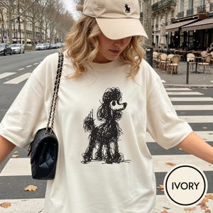 Op de afbeelding: Ivoor kleurig t-shirt met een zwarte doodle-stijl illustratie van een poedel. Het shirt heeft korte mouwen en een ronde hals. Het woord "IVORY" is in een zwarte cirkel rechtsonder gedrukt.