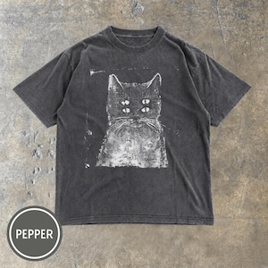 May include: A charcoal grey short-sleeve t-shirt featuring a white screen-printed graphic of a cat with four eyes. The shirt has a crew neckline and a distressed, washed-out appearance. A circular badge in the bottom left corner displays the word PEPPER.