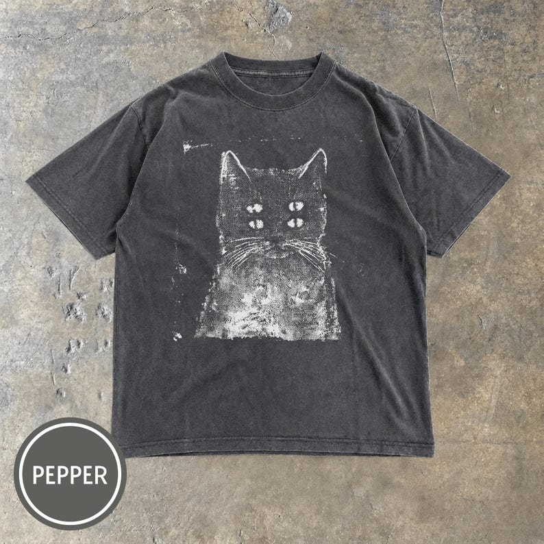May include: A charcoal grey short-sleeve t-shirt featuring a white screen-printed graphic of a cat with four eyes. The shirt has a crew neckline and a distressed, washed-out appearance. A circular badge in the bottom left corner displays the word PEPPER.