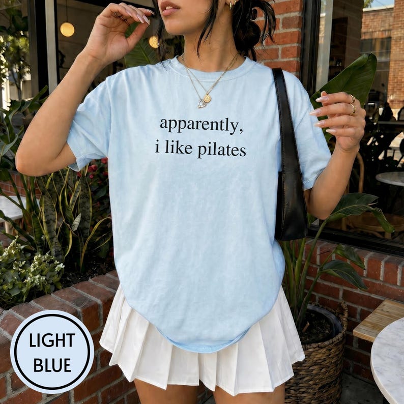 May include: Light blue t-shirt with the text "apparently, i like pilates" in black. The shirt is paired with a white pleated skirt. The image also shows a black shoulder bag and gold necklaces.