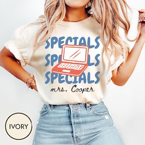 Peut inclure: Un t-shirt à manches courtes ivoire avec le mot SPECIALS en police bleue ondulée répété trois fois. Un graphique d'ordinateur portable rouge est centré entre le texte et le nom mrs. Cooper est imprimé en écriture cursive noire sous le motif.