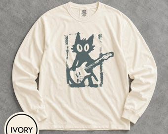 May include: An ivory long-sleeve t-shirt featuring a dark teal graphic print of a cartoon cat playing an electric guitar. The cat is centered within a distressed rectangular frame. The shirt has a crew neckline and a Comfort Colors brand label.